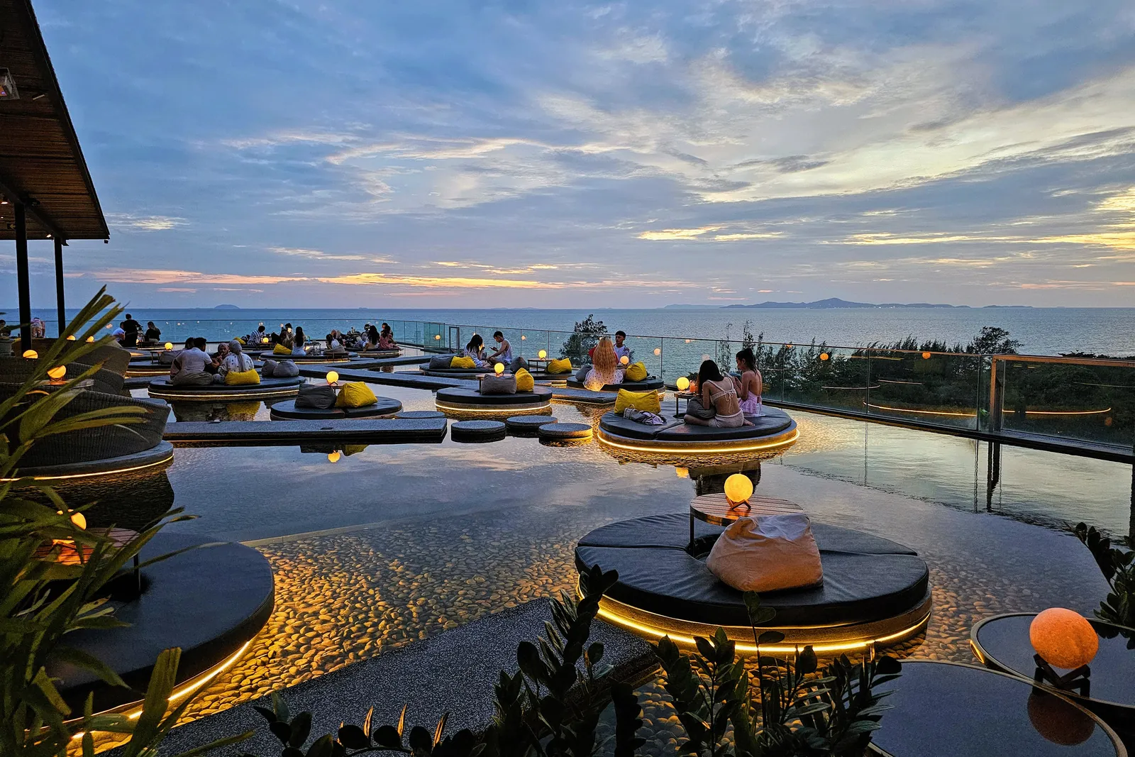 Guests on circular seating pods above the water at the Over the Moon rooftop lounge, lit by globe lamps at sunset over the sea