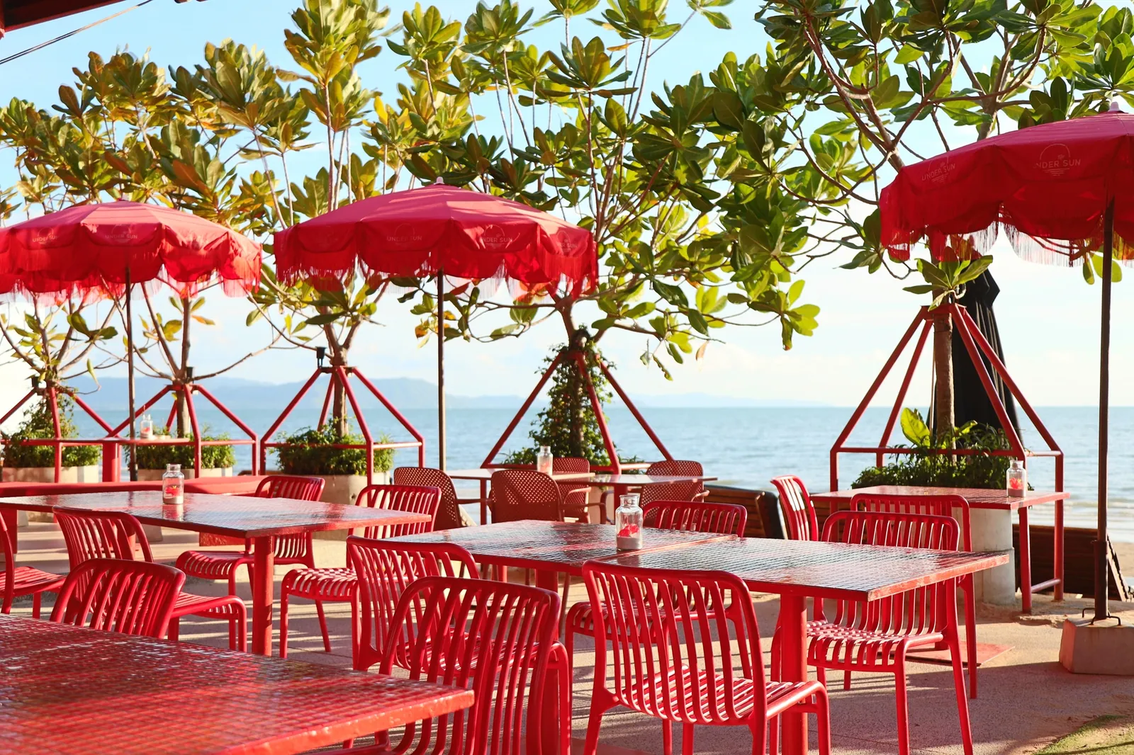 Red tables, chairs and fringed parasols on the beachfront dining terrace at Under Sun, looking out over the sea
